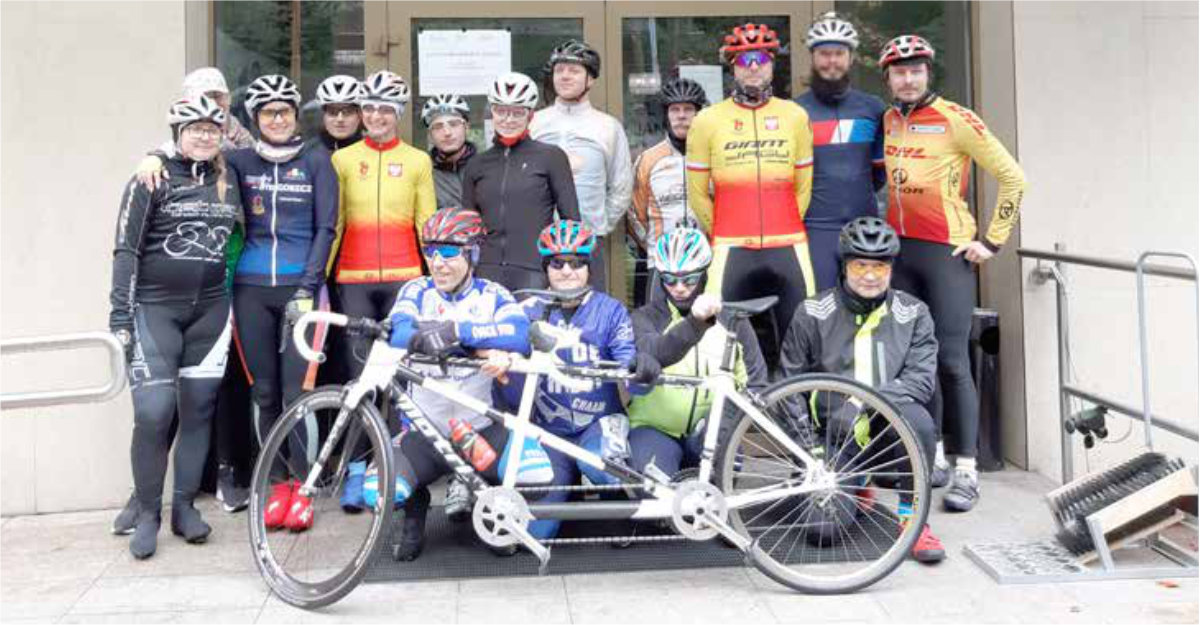 Grupa osób w strojach sportowych stojąca przed budynkiem z rowerami w tle