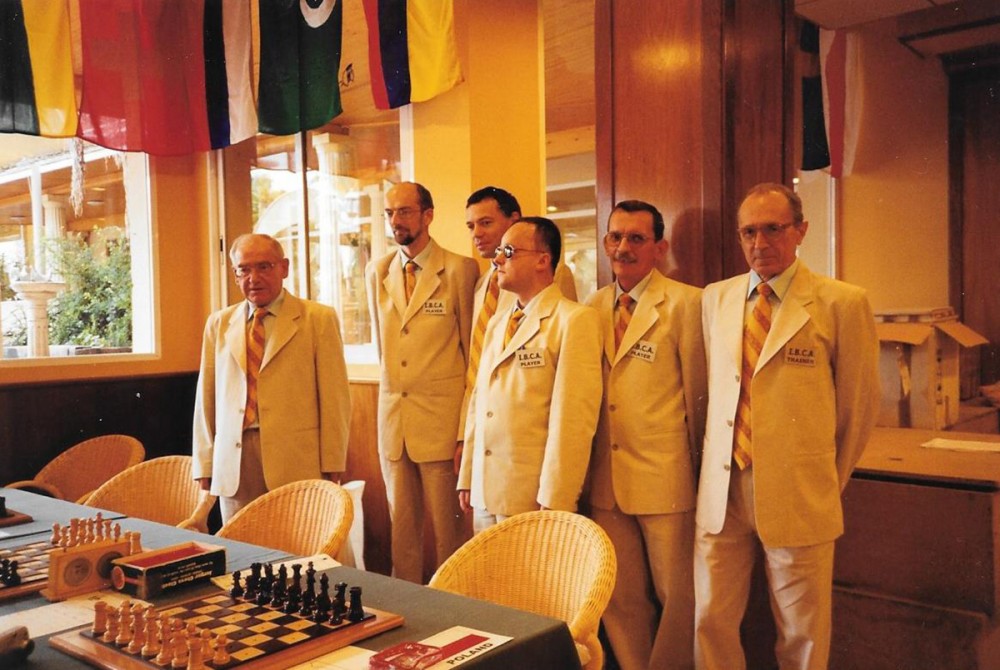 grupa osób w jasnych garniturach stojąca przed stołem z szachami