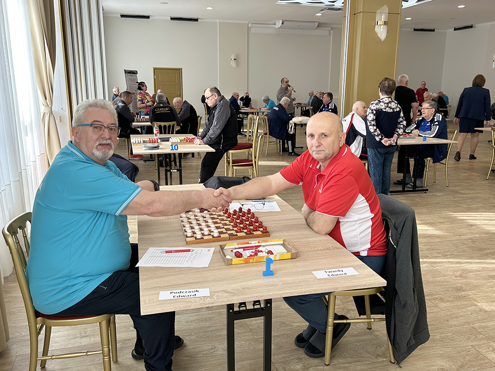 grupa osób na sali gry a na pierwszym planie dwóch mężczyzn podaje sobie ręce